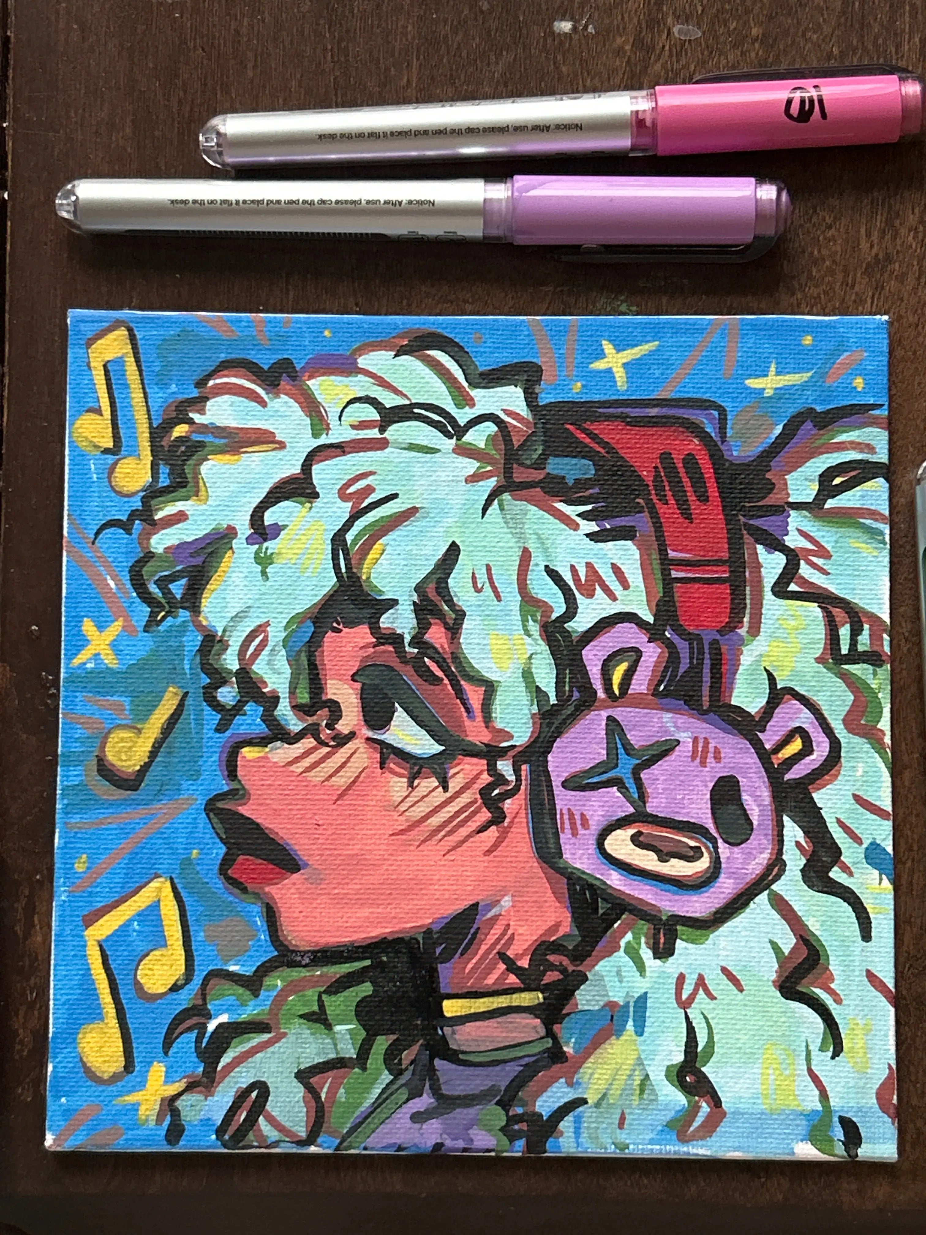 Colorful abstract painting on a small canvas with two markers on a wooden surface.