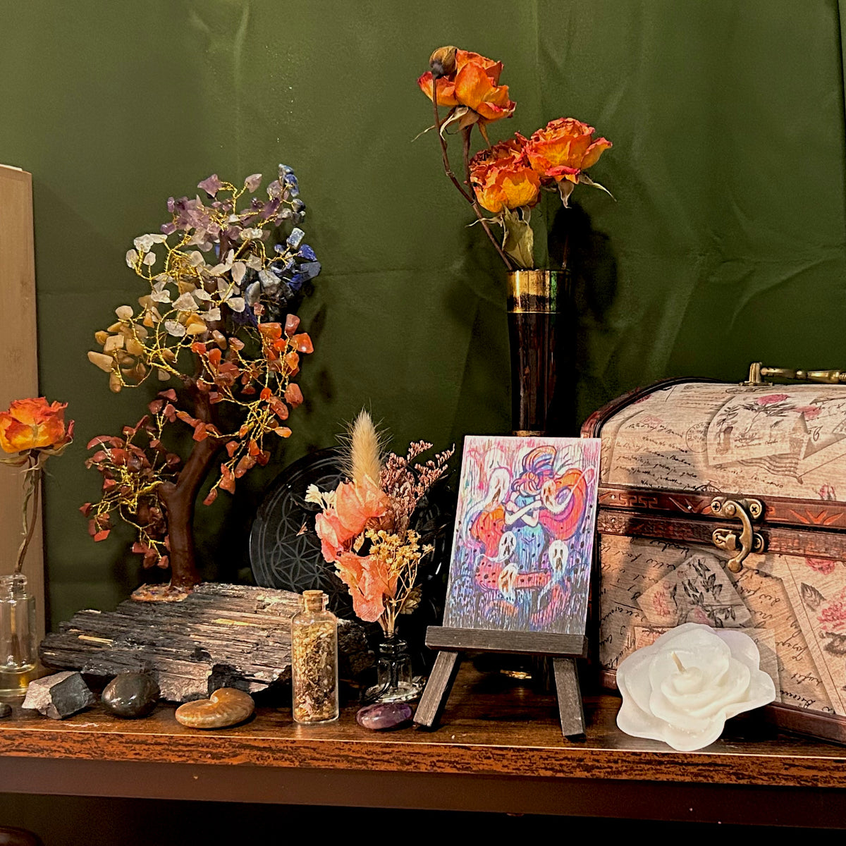 A styled tabletop display featuring a small colorful fantasy art print on a miniature easel, surrounded by dried flowers, crystals, a white rose, a vintage-style trunk, and a dark green backdrop.